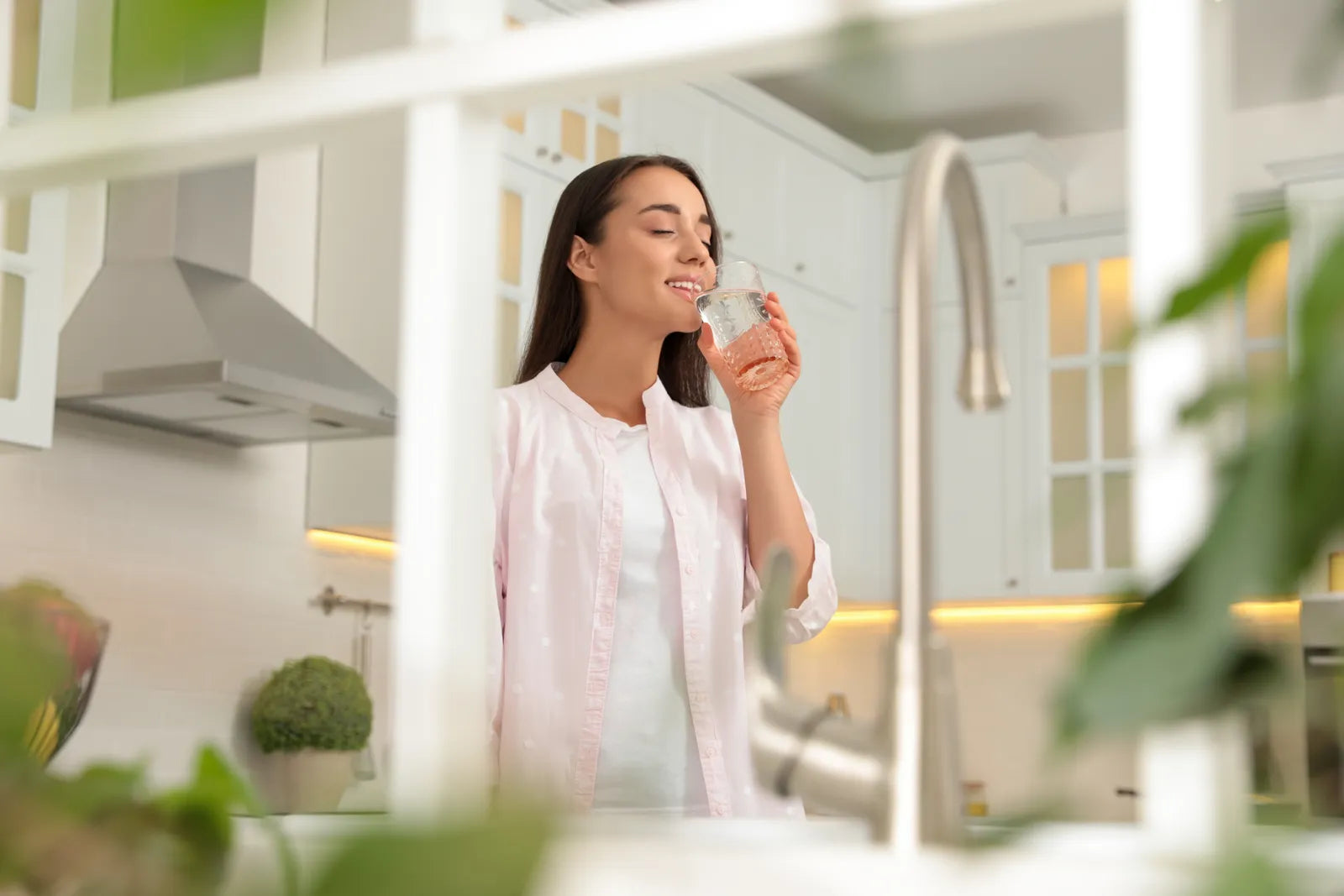 A relaxed woman drinks fresh healthy filtered water in a bright modern kitchen, enjoying clean water produced by under-sink compact reverse osmosis equipment.