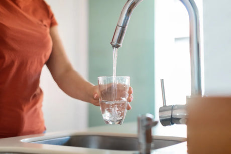 Woman holds clear glass to catch steady stream of purified water from chrome kitchen faucet connected to under-sink water filtration device.
