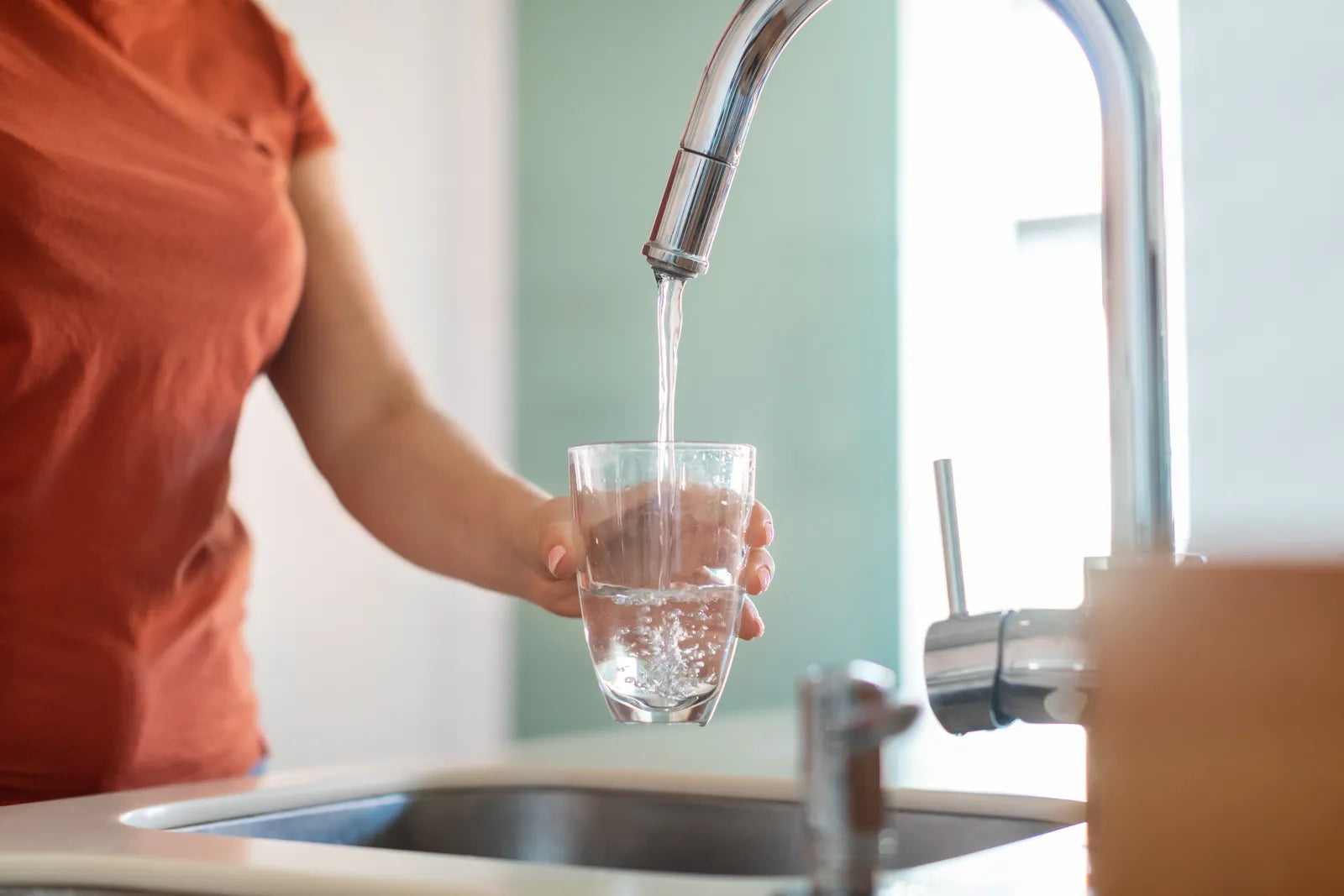 Woman holds clear glass to catch steady stream of purified water from chrome kitchen faucet connected to under-sink water filtration device.