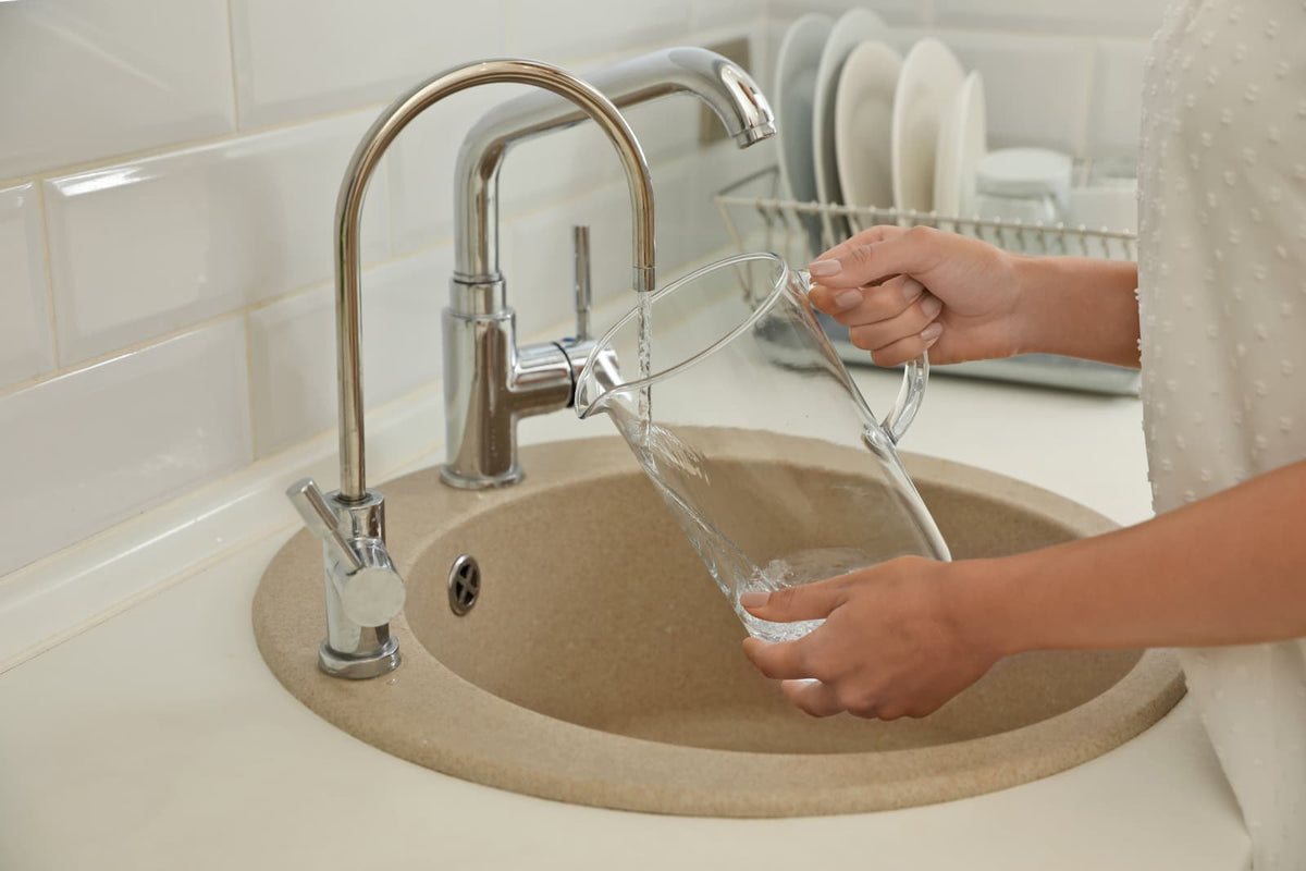 Water flows from a secondary drinking water faucet into a clear pitcher beside the main faucet