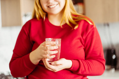 A smiling woman in a red shirt holds a glass of alkaline water in a kitchen, promoting hydration for weight loss support.