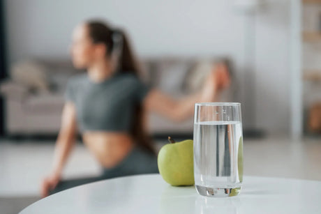 Glass of filtered water with a green apple next to a woman doing yoga, showcasing kidney-friendly hydration for active lifestyles.