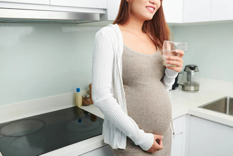 Pregnant woman holding a glass of water in kitchen, staying hydrated for a healthy pregnancy.