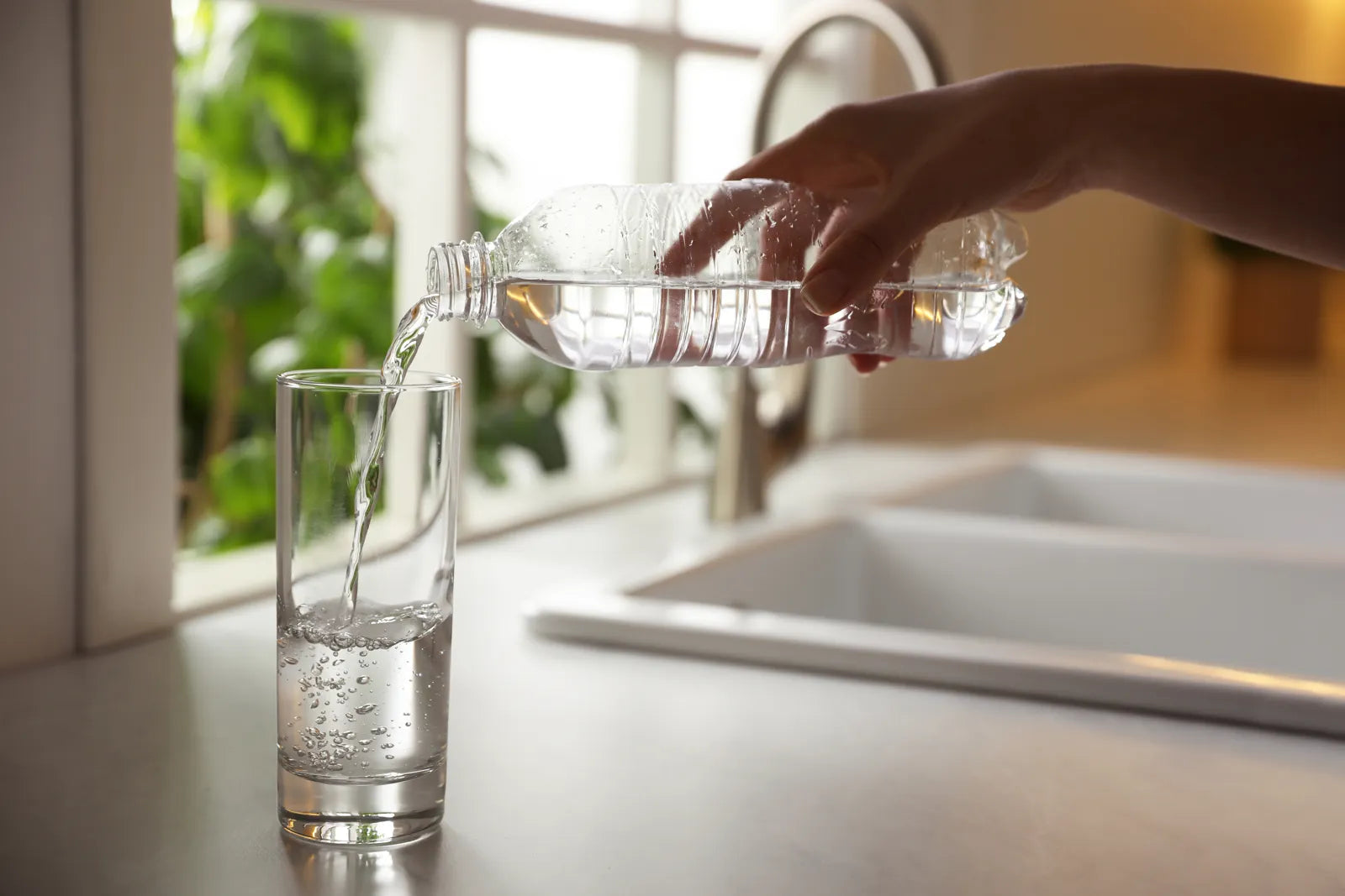 A person pours bottled water into a clear glass at the kitchen sink.