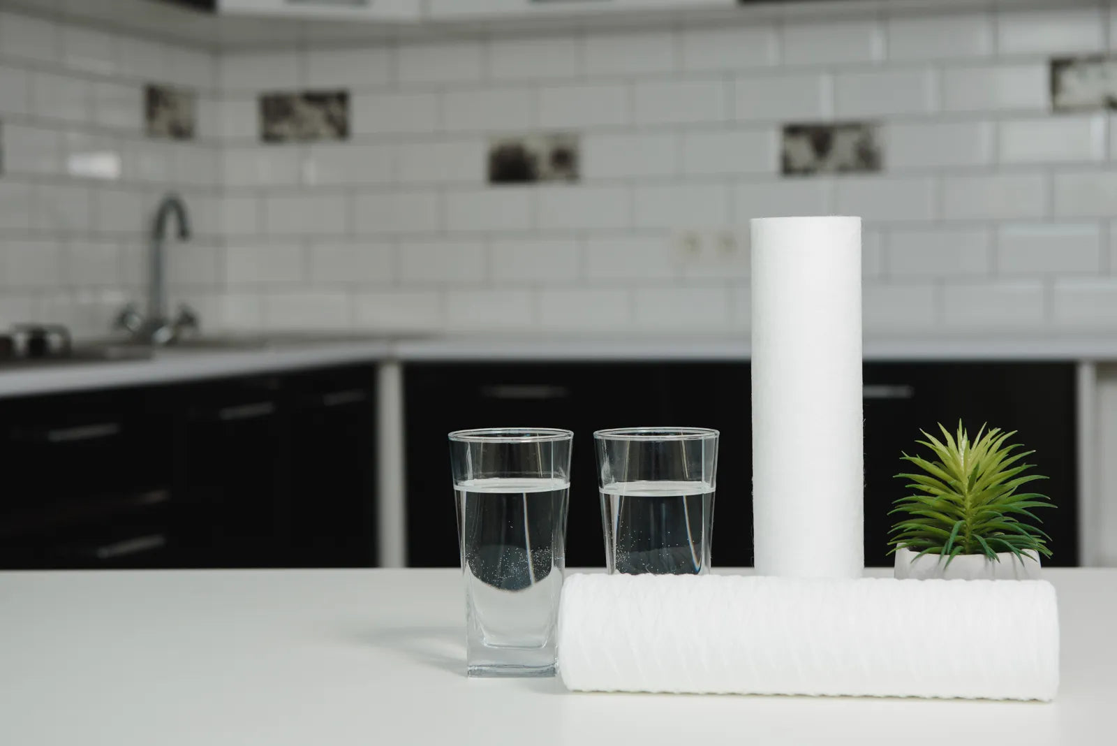 Two new water filter cartridges and glasses of water sit on a kitchen countertop, ready for replacement.