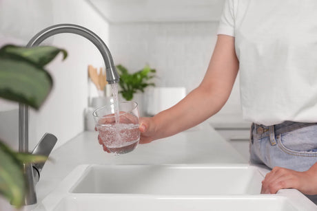 Person in white T-shirt filling a glass with clean tap water at a bright, tidy home kitchen sink.