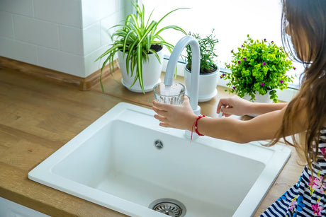 Woman fills a drinking glass from a modern kitchen faucet beside a bright white sink.