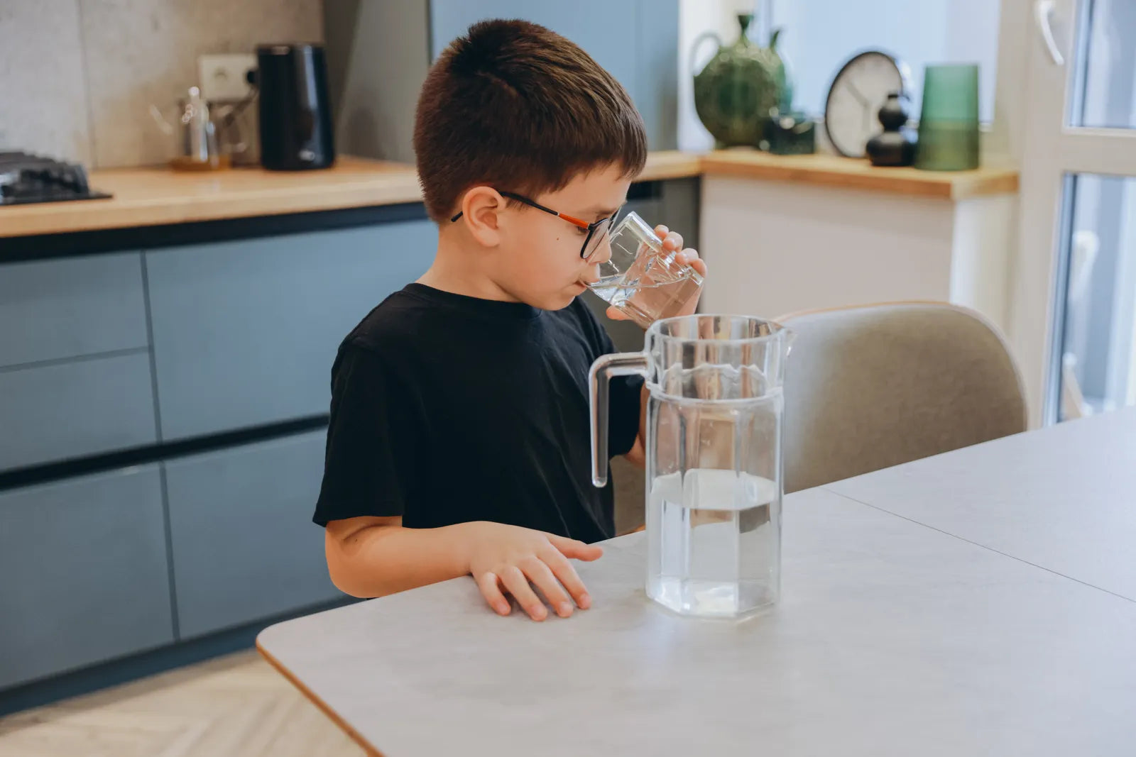 Young boy drinking filtered water from a glass beside a clear pitcher in a home kitchen, showing the use of water filters for safe, clean drinking water for children.
