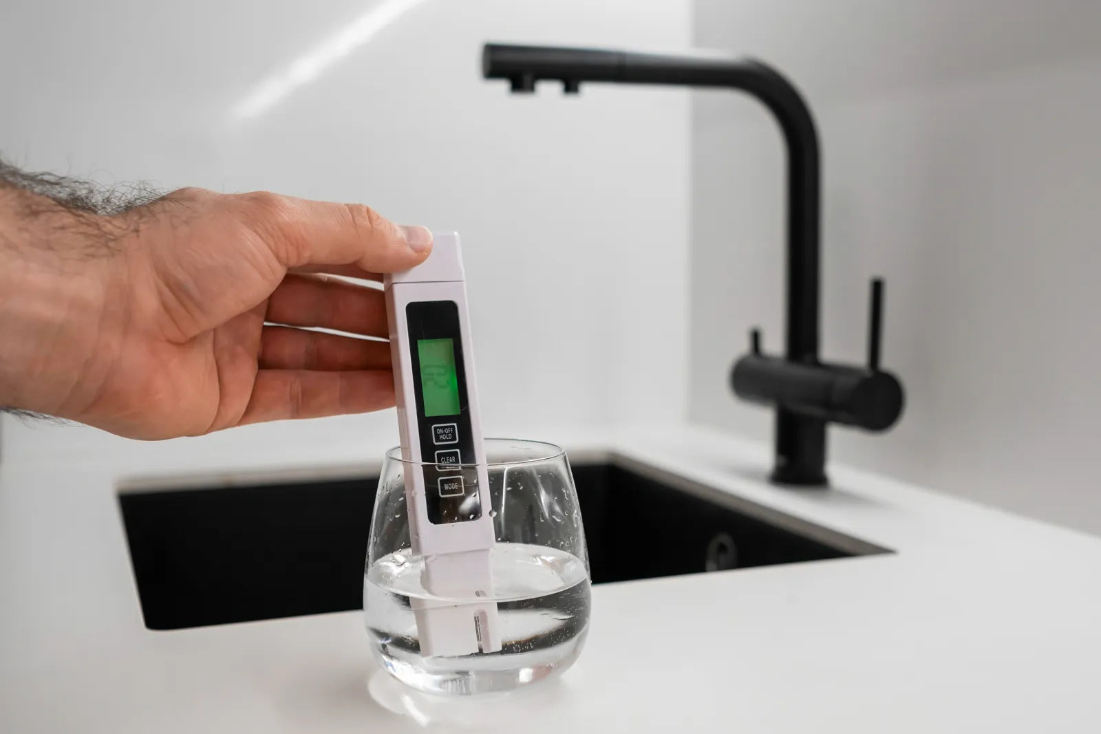 Hand using a digital TDS meter to test drinking water quality at a kitchen sink.