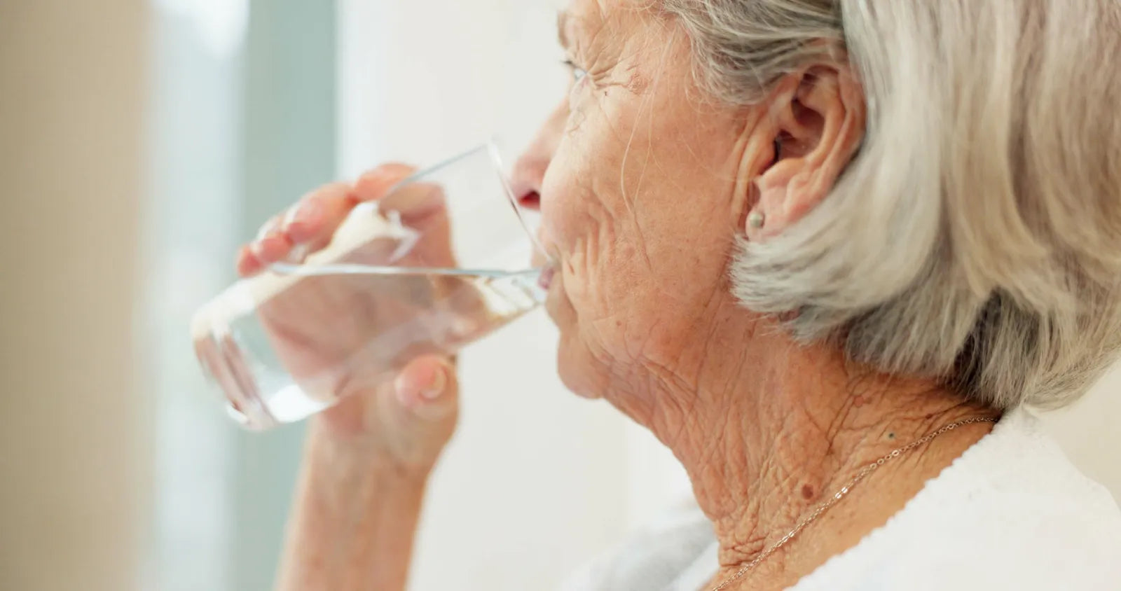 An elderly woman with gray hair drinks a glass of water indoors, supporting healthy hydration for senior well-being.