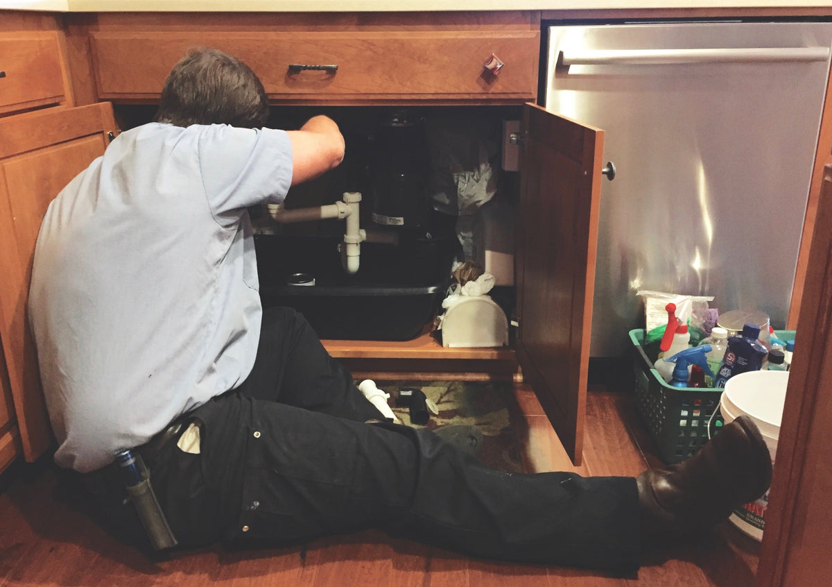 A person inspects plumbing connections and drain pipes inside an open kitchen sink cabinet.