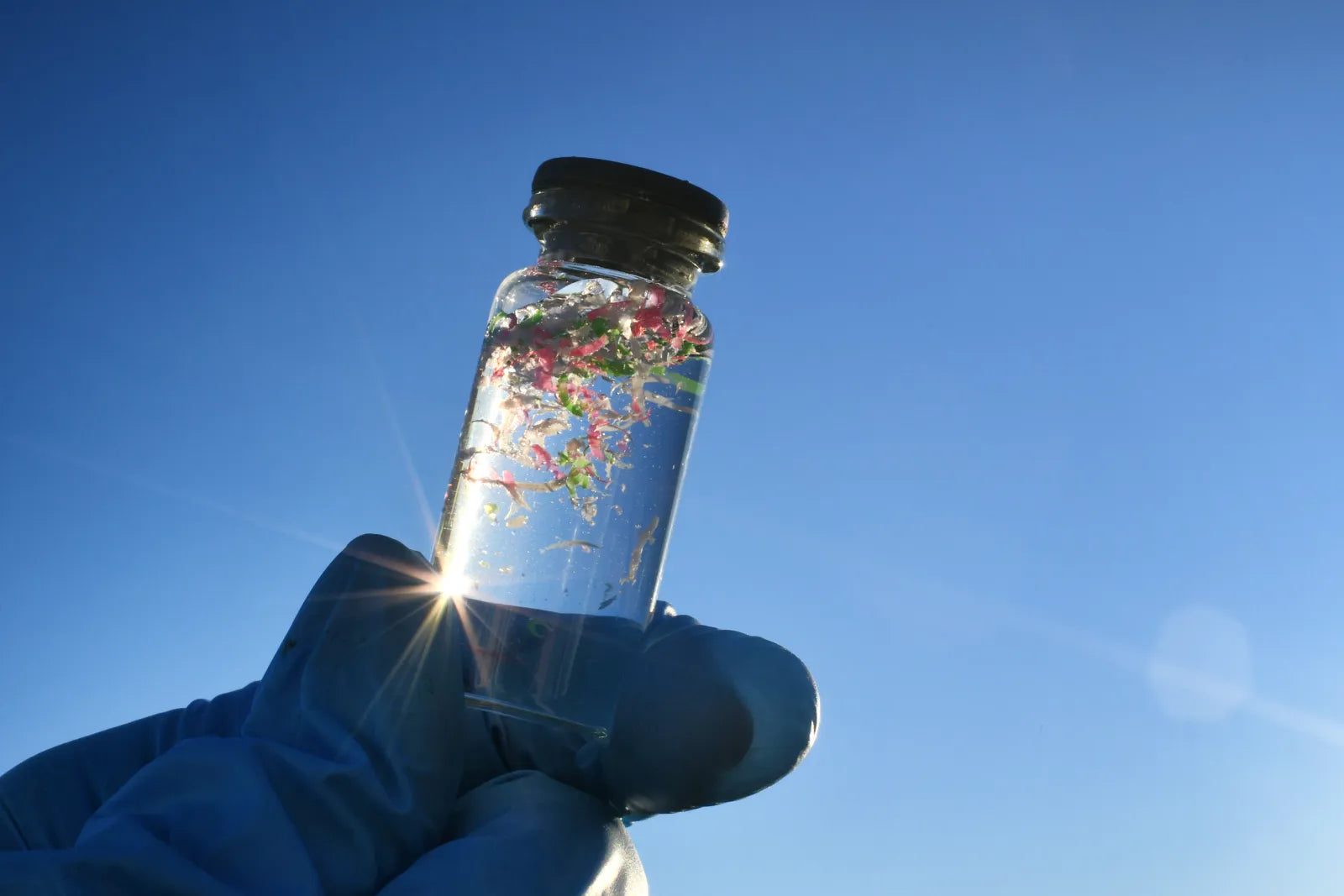 Small glass vial held by a gloved hand, containing colorful microplastic fragments in water, used to demonstrate microplastic contamination in drinking water sources.