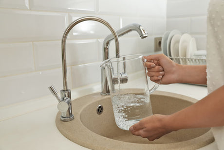 Person filling a clear glass pitcher with water from a dedicated drinking water faucet in a kitchen sink