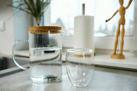Clear glass pitcher filled with water beside an empty drinking glass on a bright kitchen countertop
