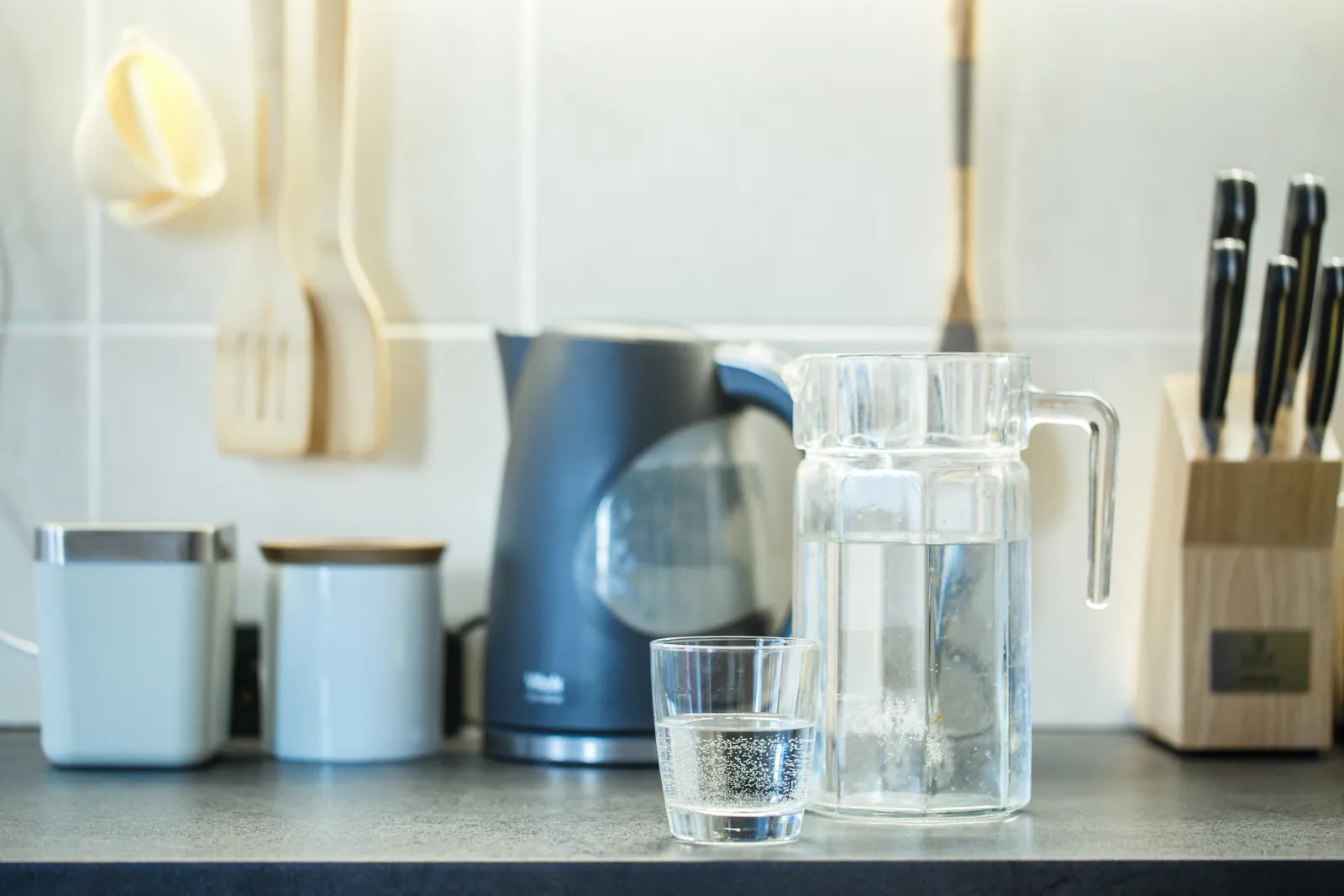 A clean kitchen counter with a glass pitcher, cup, kettle, and utensils ready for filtered water.