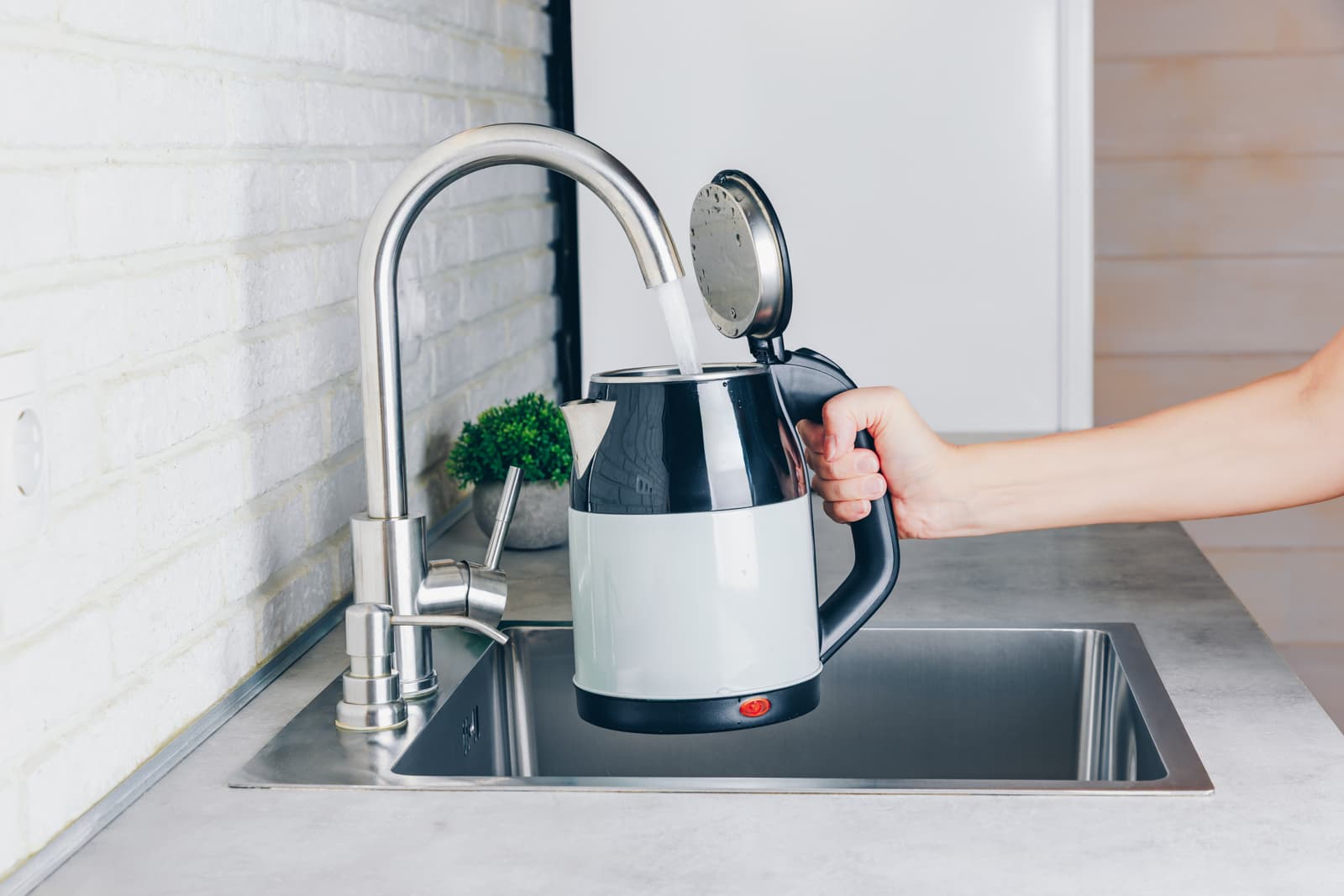 Kitchen faucet fills an electric kettle with water at a stainless steel sink for heating.
