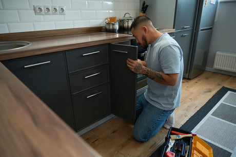 Man inspecting an under-sink cabinet while preparing plumbing for a refrigerator water connection.