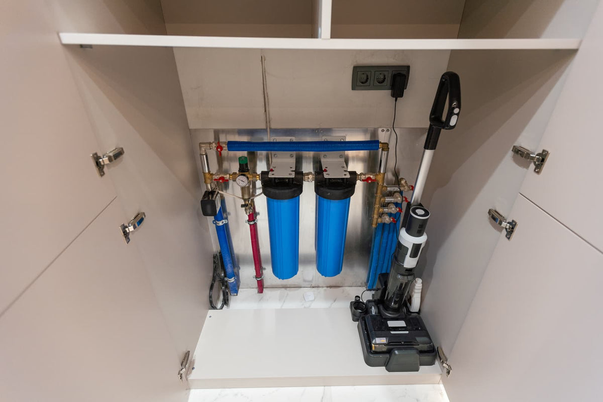Under-sink water filtration setup with blue housings, valves, plumbing lines, and nearby power outlets.