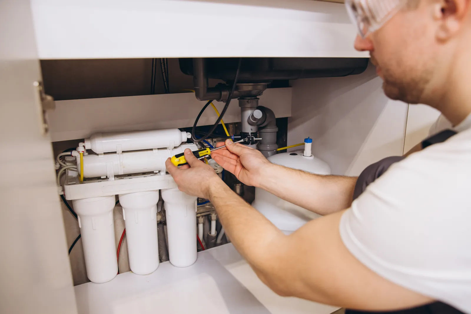 Technician uses a screwdriver to install a full-size tanked under-sink reverse osmosis filtration system inside a residential kitchen cabinet.